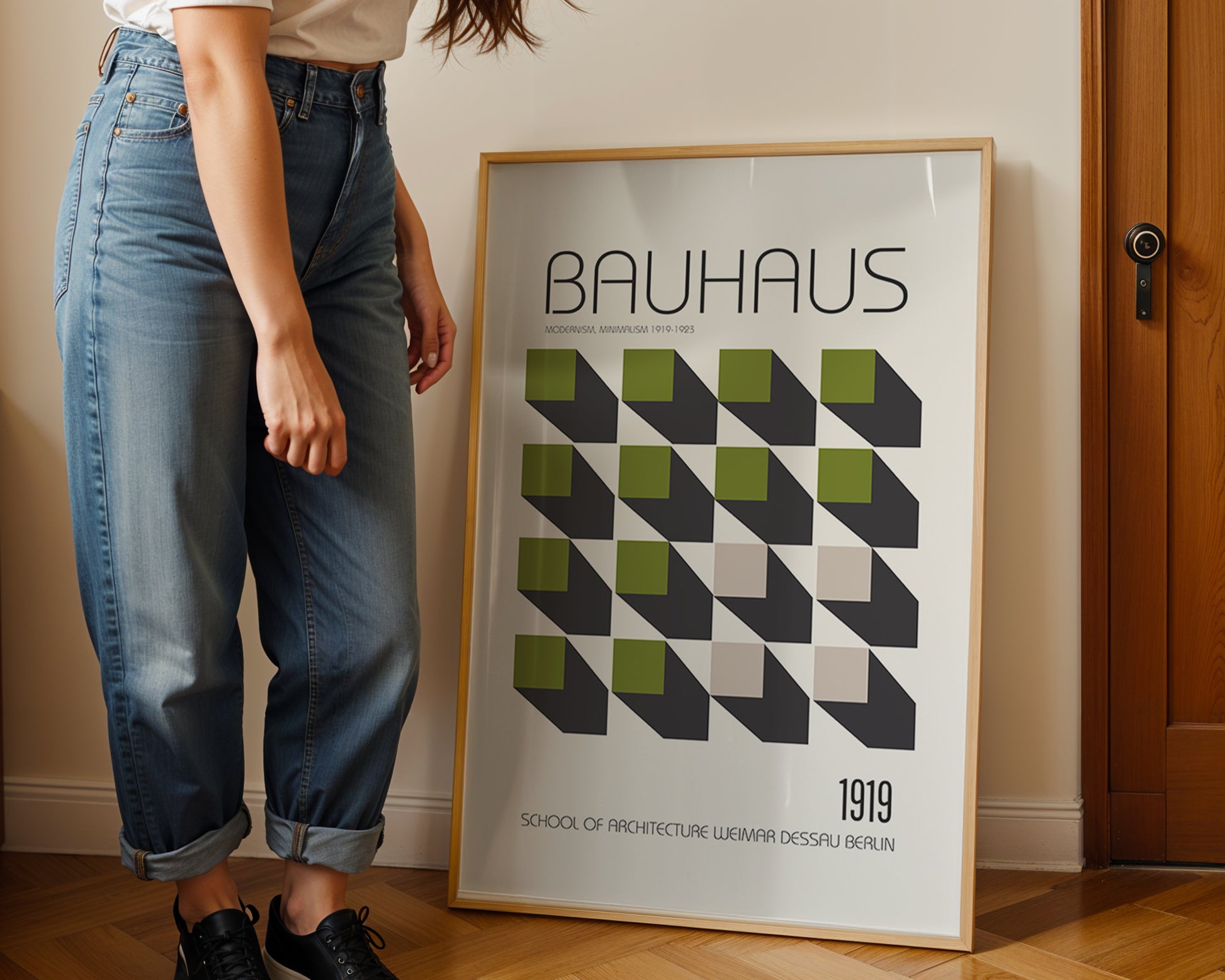 Person standing next to a Bauhaus poster in a room with wooden flooring and a door.