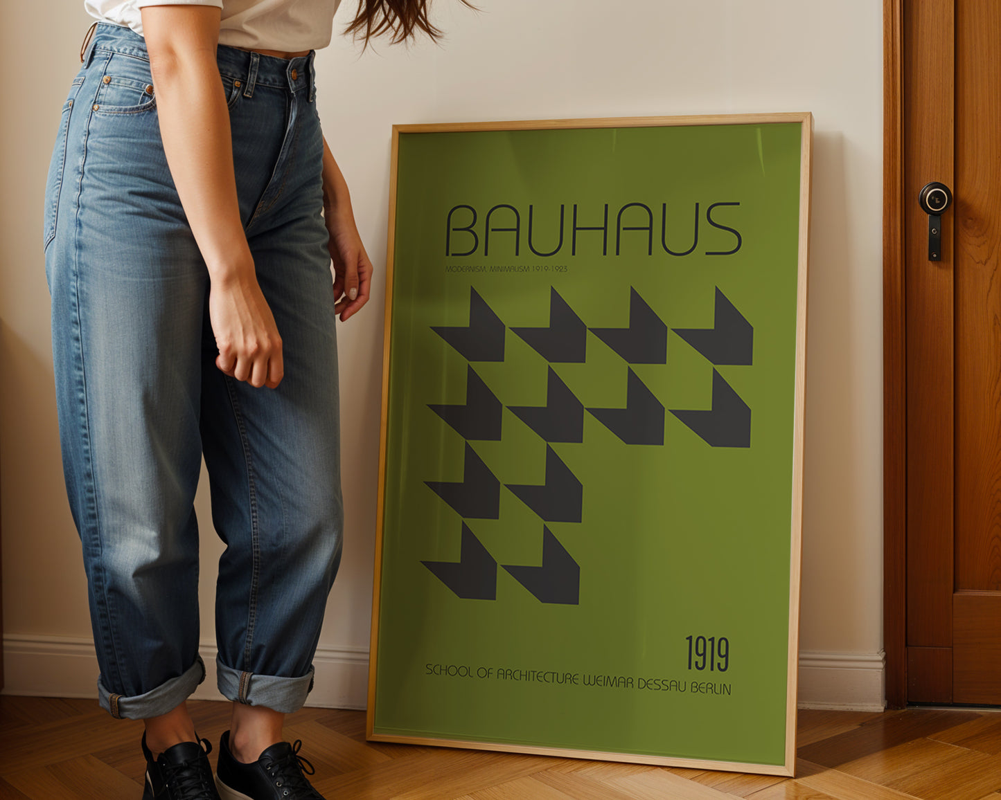 Person standing next to a Bauhaus poster in a room with wooden flooring and a door.