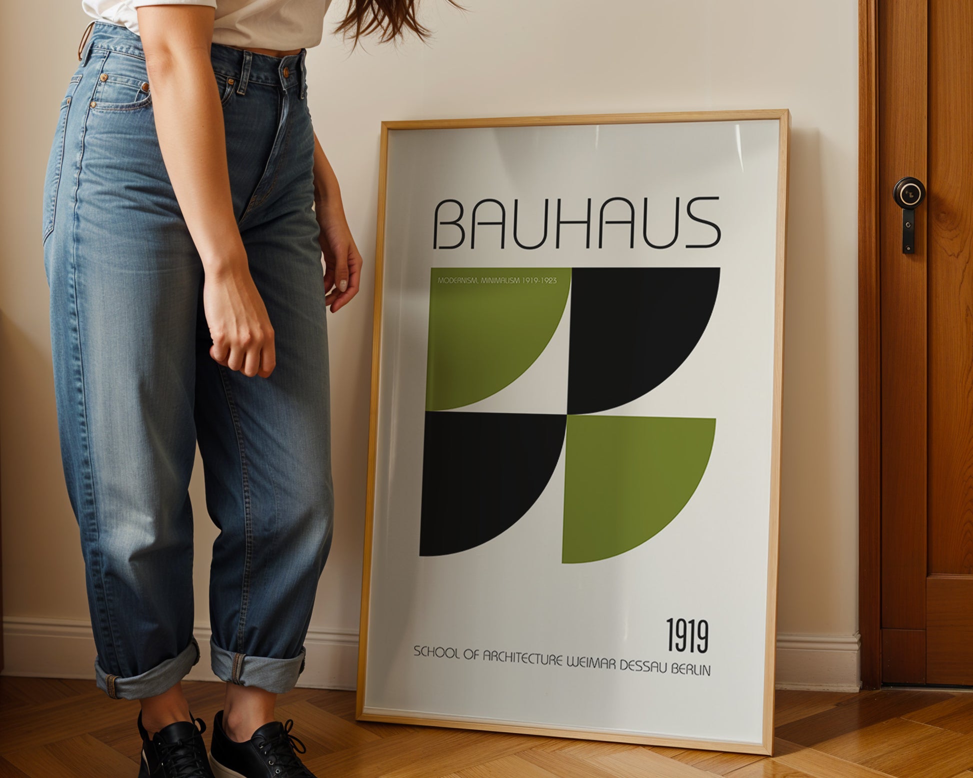 Person standing next to a Bauhaus poster in a room with wooden flooring and a door.