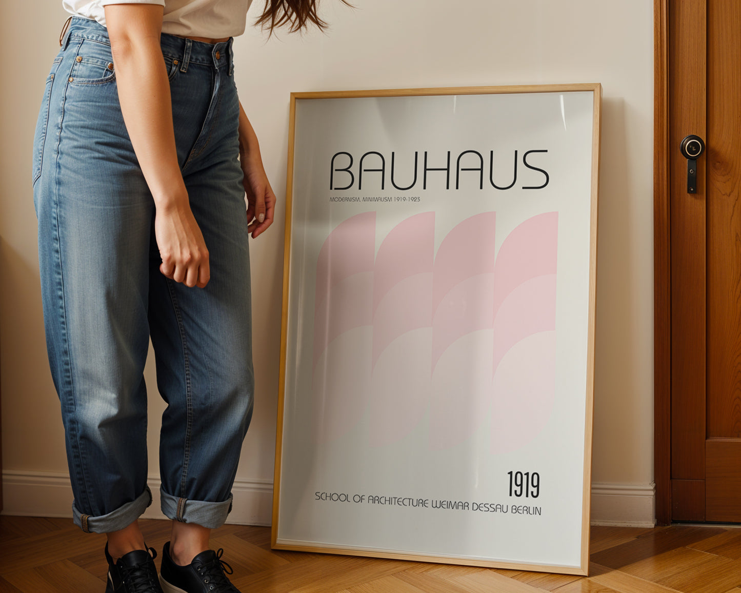 Person standing next to a Bauhaus poster in a room with wooden flooring and a door.