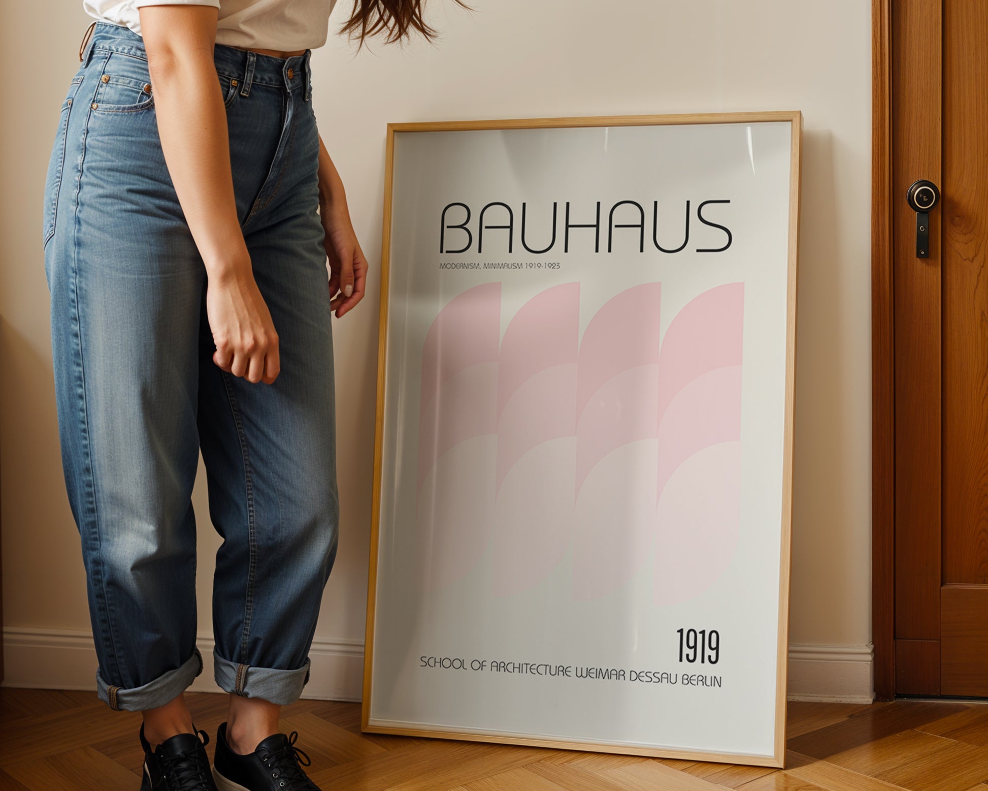 Person standing next to a Bauhaus poster in a room with wooden flooring and a door.