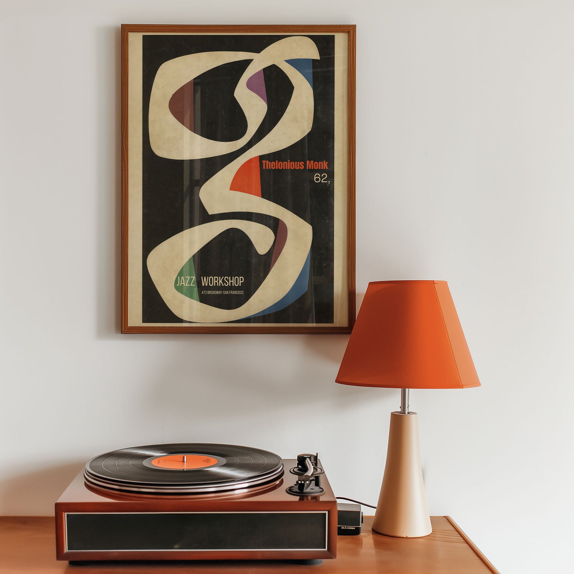 Wooden record player with a vinyl record on a wooden table, framed abstract art above, and an orange lamp to the right.